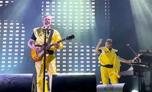 Two men in yellow jumpsuits perform on stage, one playing guitar and the other at a keyboard.