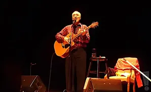 Singer-songwriter plays an acoustic guitar and sings into a microphone on stage.
