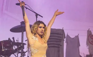 Singer with arms raised, holding a microphone on a stage with a purple background and laundry hanging.