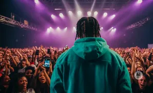 A performer in a teal hoodie faces a large, cheering crowd under bright purple stage lights.