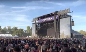 A large outdoor concert crowd watches a band perform on a stage that reads 