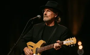 Musician wearing a black hat and suit plays a tan guitar on stage.