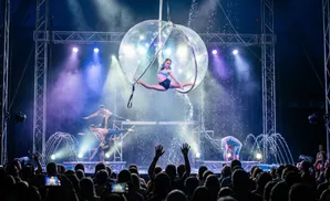 Acrobats perform in a water-filled sphere and on stages with fountains, while a crowd watches.