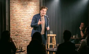 A comedian performs in a dimly lit club to an audience sitting at tables.
