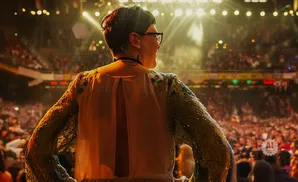 Woman with short dark hair and glasses on stage, looking out at a cheering crowd under bright lights.