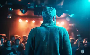Man in a teal sweater facing away from the camera, on a stage in front of a crowd.