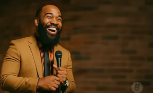 A Black man with a beard in a tan suit laughs into a microphone in front of a brick wall.