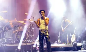 A singer in a white tank top and shiny black pants performs on stage with a band.