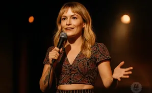 Woman with blonde bangs and a patterned crop top holds a microphone, gesturing with her hands.