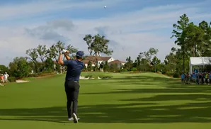 Golfer hits ball on a sunny day with spectators and a house in the background.