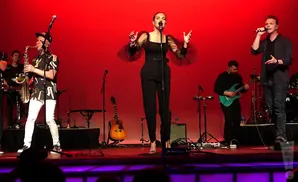Band performing on a stage with red backdrop. A woman in black sings center, with a saxophonist left and a guitarist right.