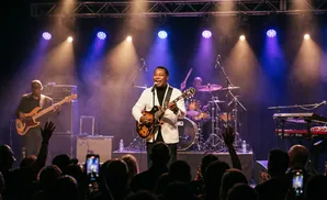 A band performs on stage with purple and yellow spotlights, a singer in a white jacket plays guitar.