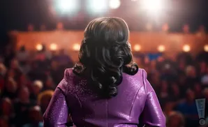 A woman with dark, curled hair in a sparkly purple jacket faces away from the camera, on a stage in front of a blurred audience.