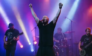 A male singer with arms raised sings into a microphone on a stage with colorful lighting.