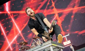 A male guitarist plays an electric guitar on a stage with a red, abstract background at a concert.