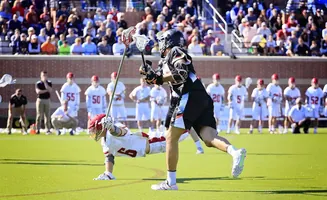 Lacrosse players compete on a green field, one on the ground, the other with a stick raised.