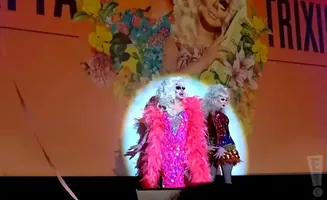 Two drag queens with platinum blonde wigs and heavy makeup perform on stage. The queen on the left wears a sparkly pink dress and a pink feather boa. Th…