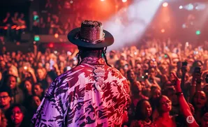 Rapper in a hat and floral shirt on stage with a crowd cheering and holding up phones.