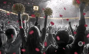 People with pom-poms cheer in a crowd with pink confetti falling.