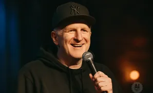 A man in a black baseball cap and hoodie smiles while holding a microphone.