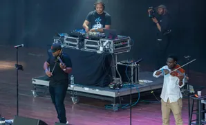 A DJ and two violinists perform on stage with audio equipment.