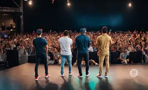 Four men stand on a stage facing a large, cheering crowd with their phones up.
