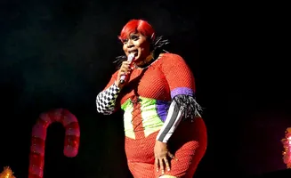 A plus-size performer in a red, sequined outfit and red wig sings into a microphone on a stage.