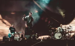A band performs on a dimly lit stage with a large screen behind them. The lead singer, in a tracksuit, sings into a microphone.