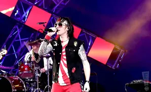 A rock singer with sunglasses and tattoos performs on stage, wearing a denim vest over a red and white striped shirt and red shorts.