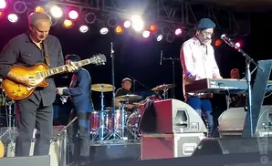 A band performs on stage with a guitarist, keyboardist, and drummer under bright lights.