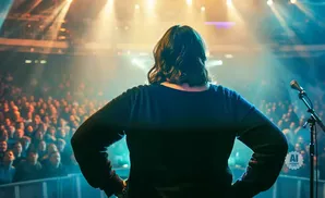 A person on stage faces a large audience under bright spotlights, with a microphone to their right.
