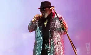 A man in a sparkly jacket and fedora sings into a microphone while holding a cane.