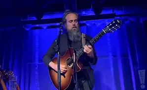 A man with a long beard plays an acoustic guitar on a blue-lit stage.