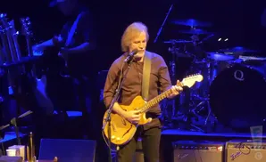 A man with long, graying hair plays a guitar on stage. He wears a brown shirt and black pants.