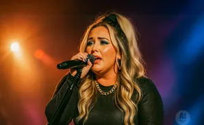 A woman with blonde hair sings into a microphone on a brightly lit stage.