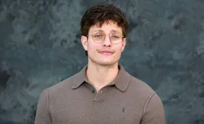 A man with glasses and a brown polo shirt smiles against a dark, textured background.