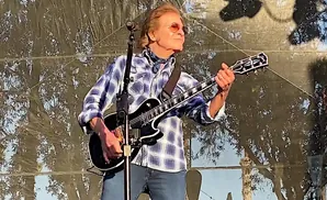 A man in a blue plaid shirt plays a black electric guitar on stage.
