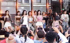 A group of young women perform on stage, with fans holding up phones to record.