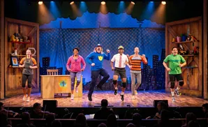 A group of actors in costumes dance on a stage with a cityscape backdrop and shelves of toys.