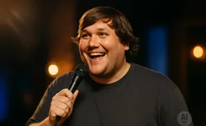 A man with dark hair, wearing a grey t-shirt, laughs while holding a microphone.