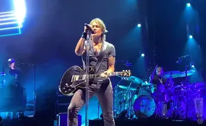 A man plays an electric guitar and sings into a microphone on a stage with blue lighting.