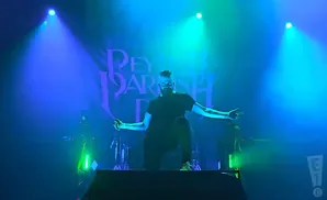 A man with a beard and a black t-shirt performs on stage with blue and green lights behind him.