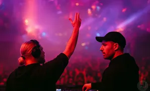 Two DJs, one with headphones and a bun, the other wearing a baseball cap, perform on a stage with pink and purple lights.