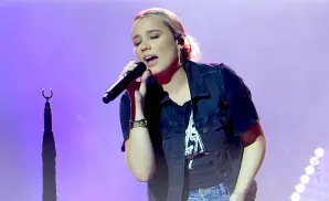 Singer Gabby Barrett performs on stage, holding a microphone and wearing a denim vest over a t-shirt.