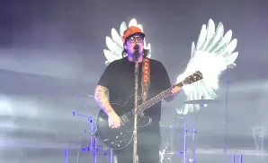 A man in a black shirt and orange hat plays an electric guitar on stage with glowing white wings behind him.