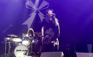 A female singer performs on stage with a drummer and a large white ribbon logo in the background.
