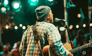 A man in a beanie and plaid shirt plays guitar on stage with a microphone in front of him.