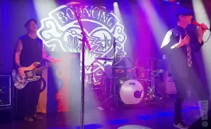A band performs on stage with purple and white lighting. The bassist is on the left, drummer in the center, and singer on the right.
