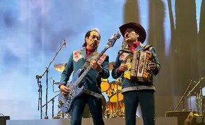Two men on stage in matching teal suits with ornate collars play music: one plays a blue bass guitar, the other an accordion.