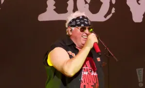 A man with a bandana and sunglasses sings into a red microphone on stage.